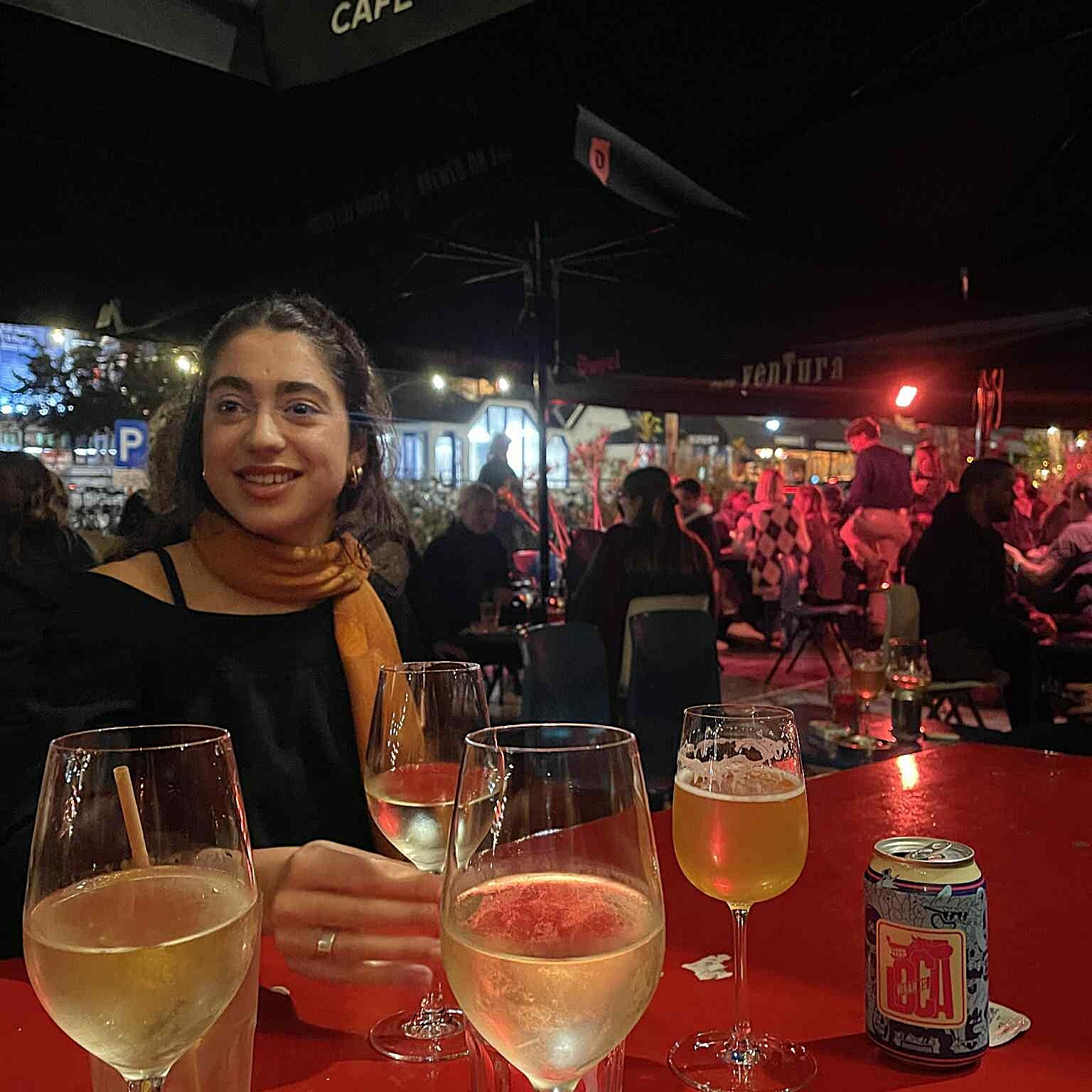 A girl with an orange scarf is sitting at a red table. She is drinking a glass of wine. On the table are two glasses of wine and a beer.