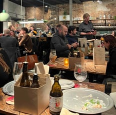 A cozy evening scene inside OTOMAT restaurant in Ghent, with people enjoying food and drinks at wooden tables in a warm, industrial-style setting.