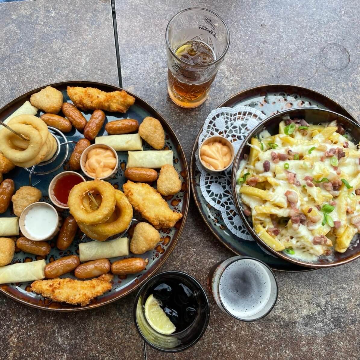 marble table with cheesy fries and a plate full of fried snacks, like springrolls, onion rings, calamaris, chicken tenders, with some drinks