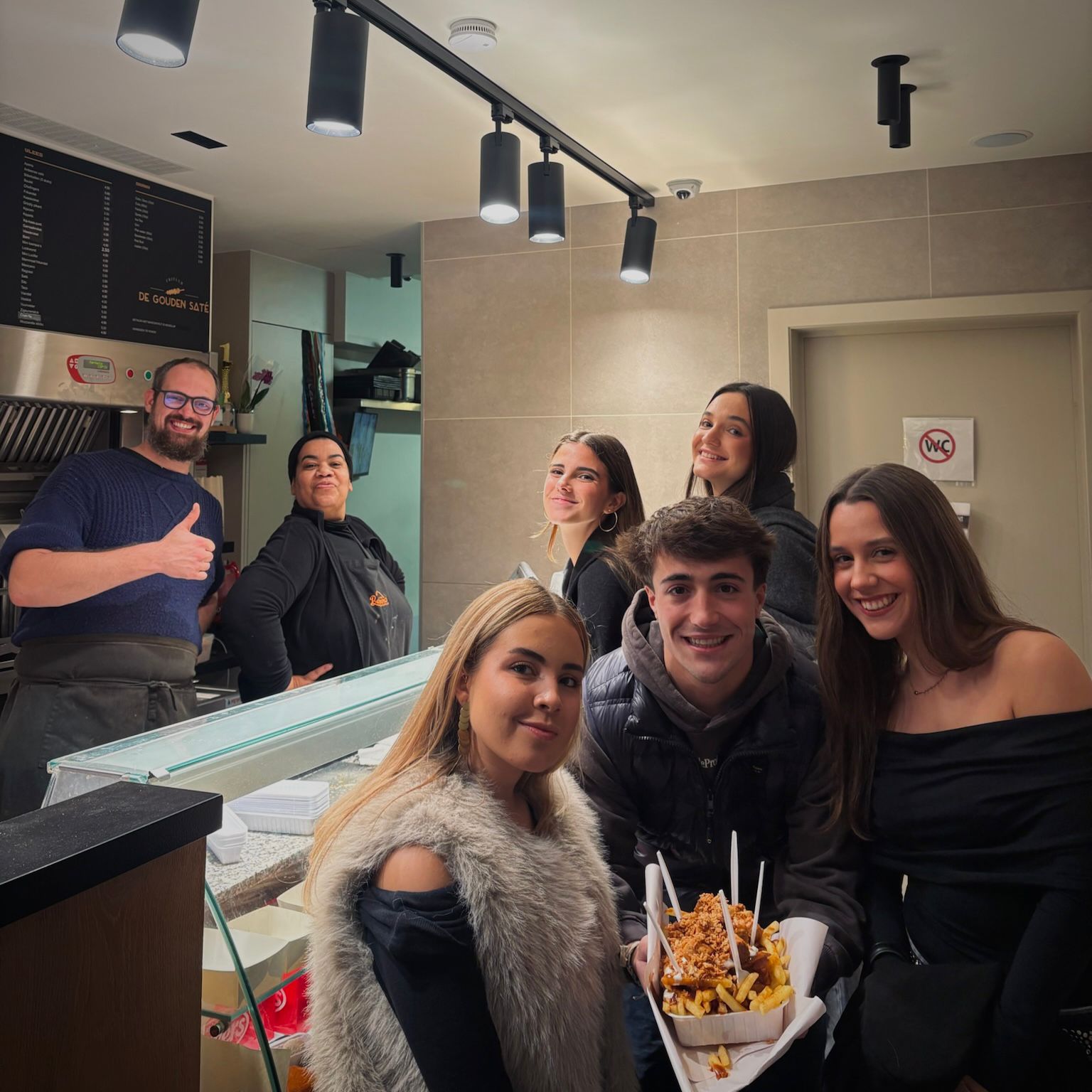 Group of students smiling with staff at a late-night snack bar in Gent, holding fries with sauce.
