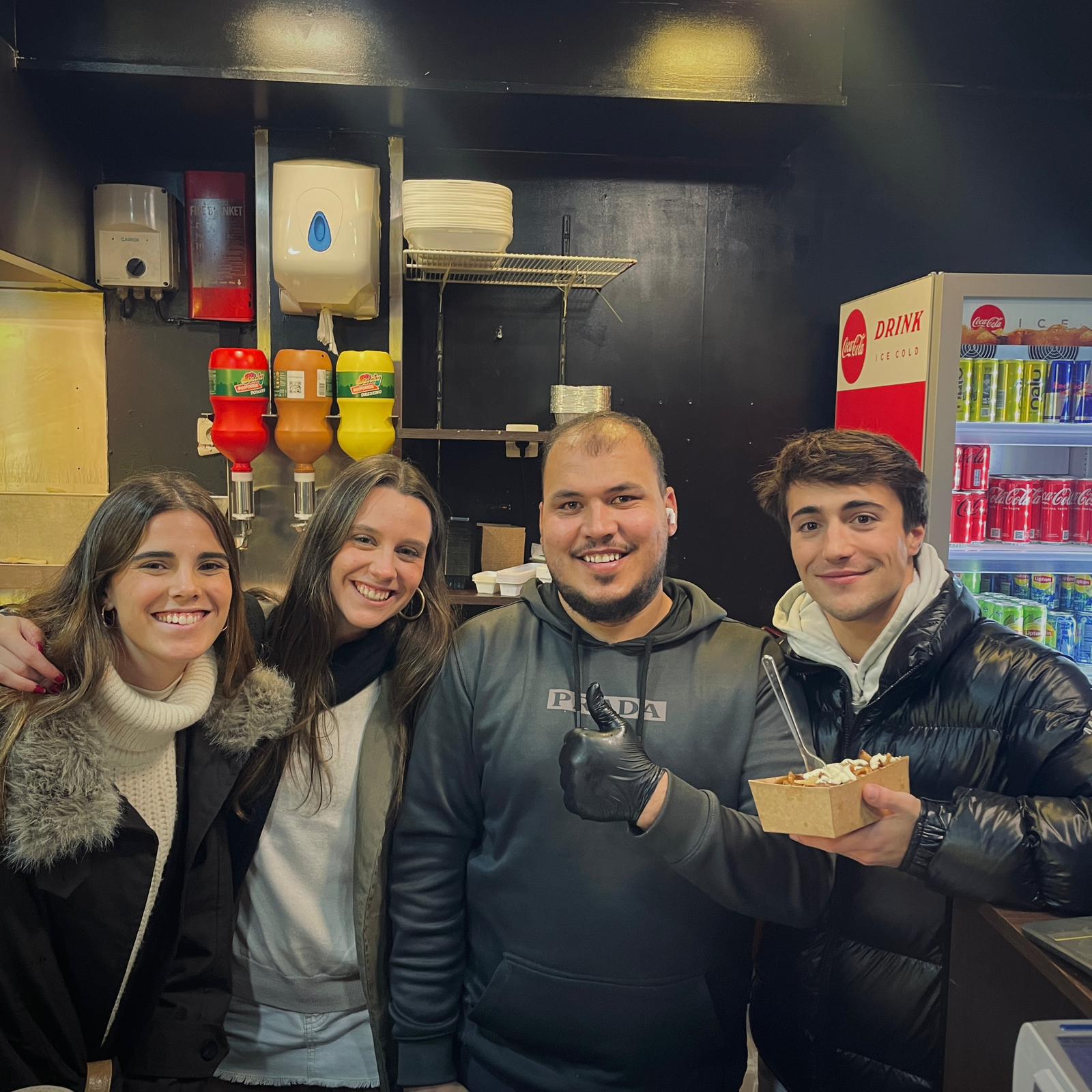 Erasmus students posing with the cook inside a Gent fast-food spot, holding a box of fries.