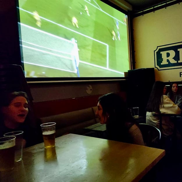 2 girls talking to each other and drinking beer while a football game is showing on a large screen behind them