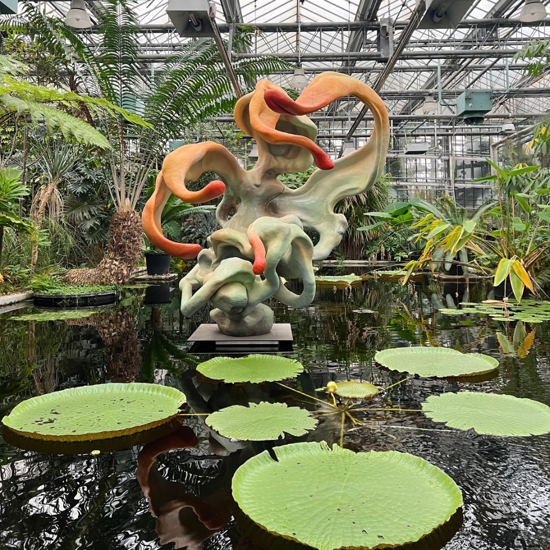 A bold green sculpture is positioned in a pond, encircled by water lilies and framed by lush tropical vegetation.