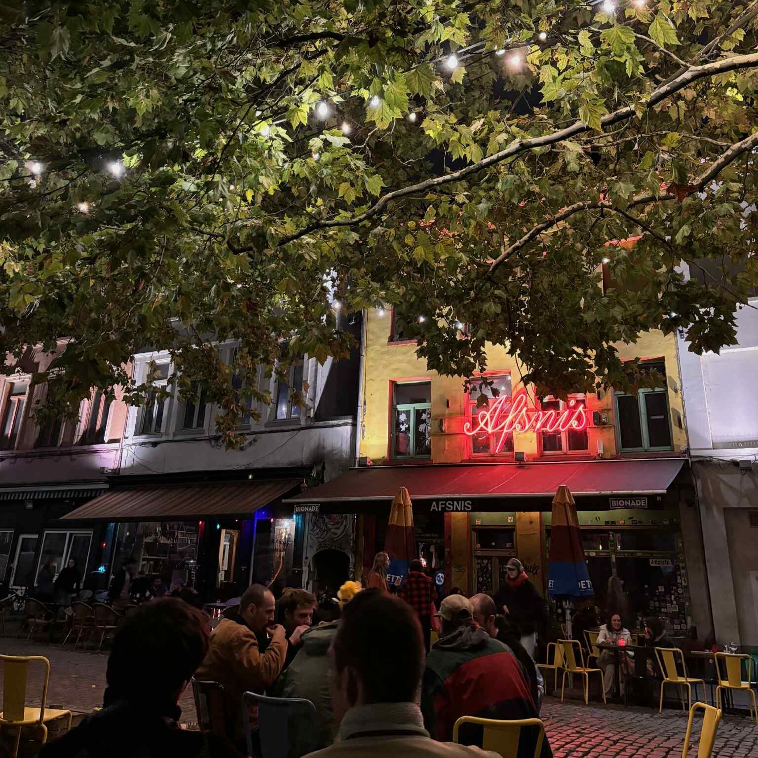 People sitting outside a bar on yellow and blue chairs. There is a red neon sign on the facade with the word 'Afsnis'.