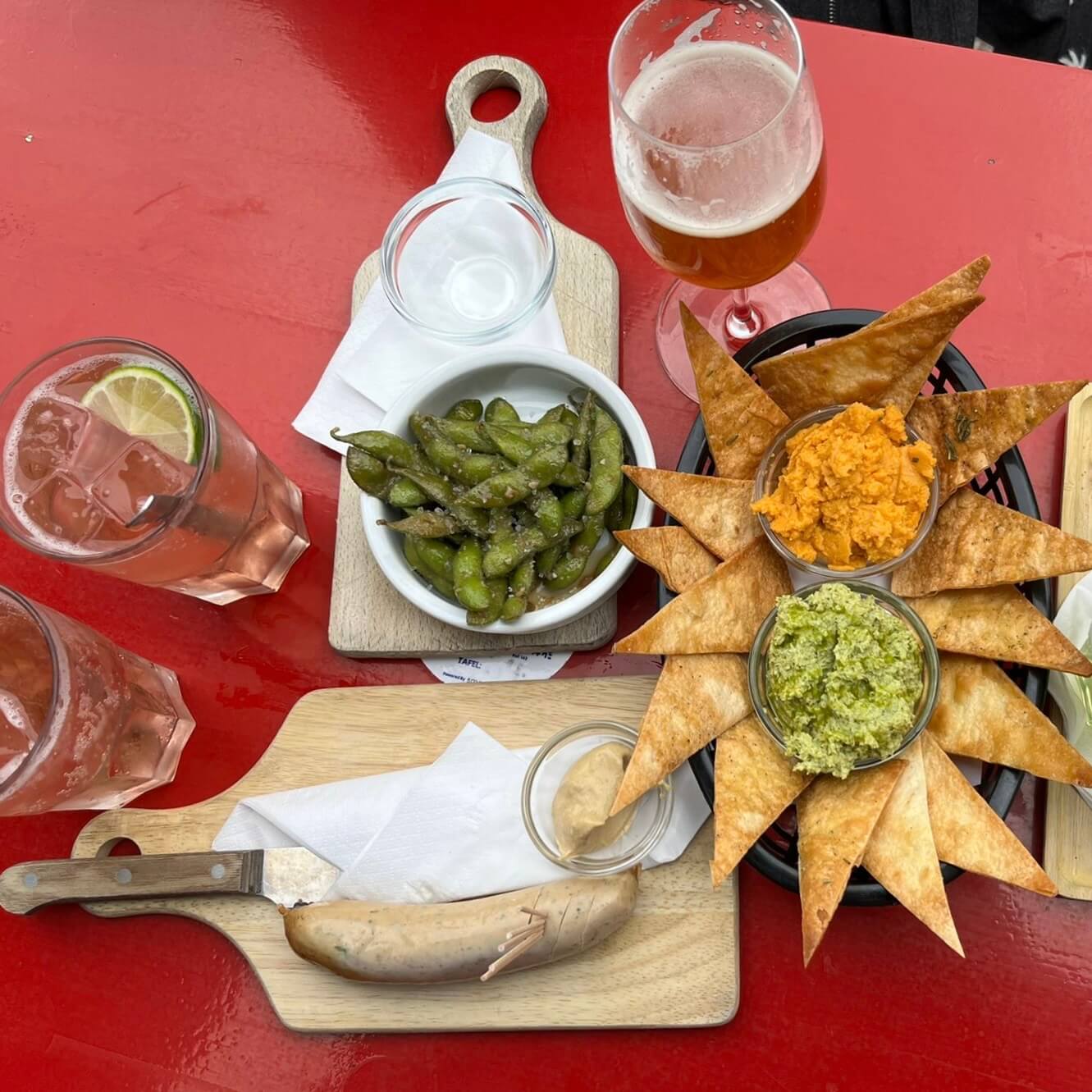 red table with edamame beans, a white pudding with mustard, two vegetarian dips with long triangular crackers, and some drinks