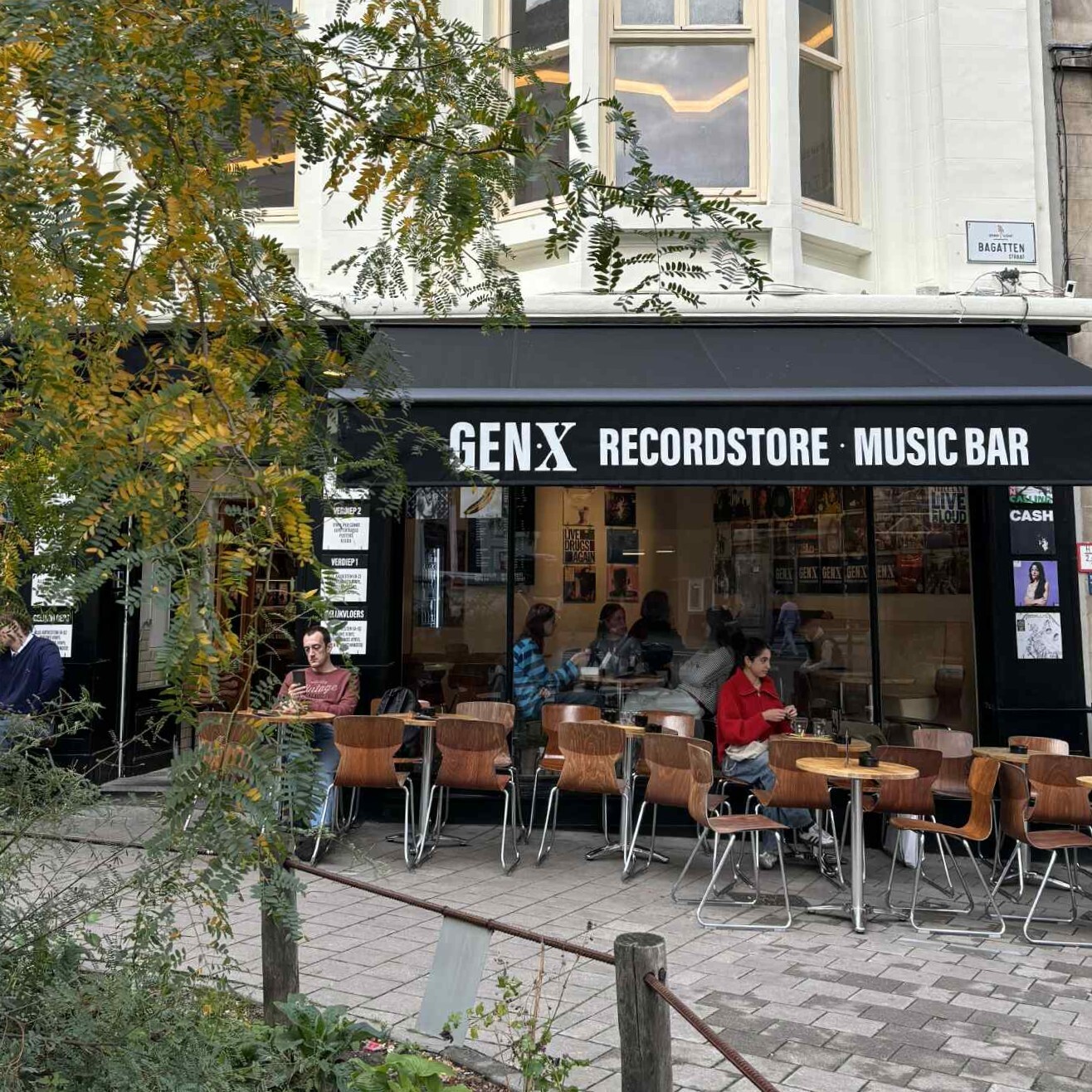 A coffeebar at the corner of the street. Outside are wooden chairs and some people who are drinking a coffee.