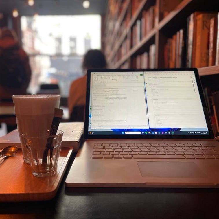 A computer and a delicious cup of coffe on one of the tables of Le bal infernal. With the used bookshelves in the background, setting a cozy and welcoming mood for your studies