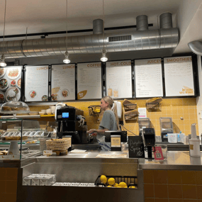 Oats Day long counter with baked goods and an employee at the cash register and menus on a yellow tile wall.