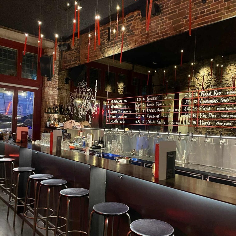 Interior of a cozy Belgian bar with a long counter, high stools, exposed brick walls, and a board displaying various beers on tap.