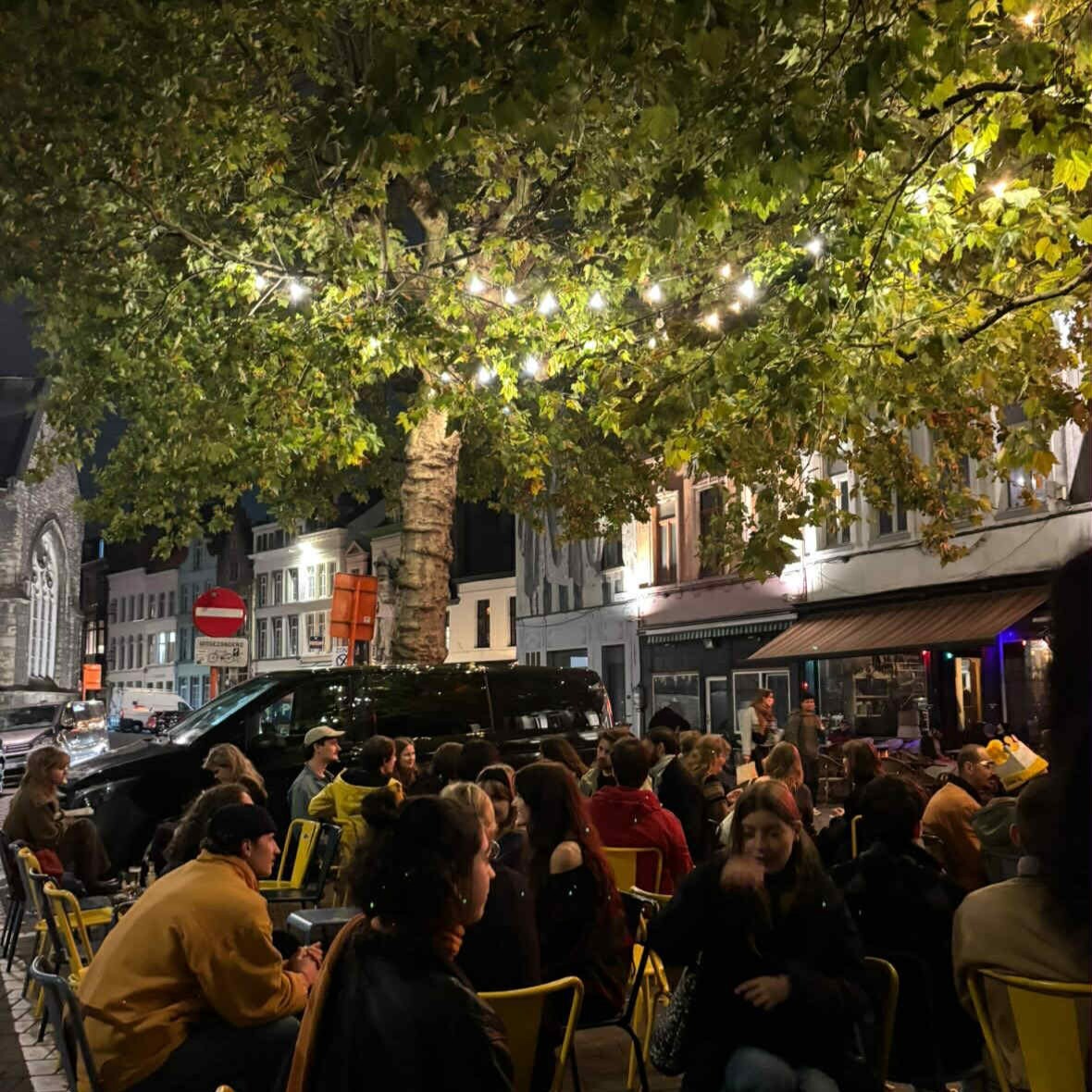 There are people sitting on yellow chairs in the streets and they are chatting. There is also a tree with lights hanging in it.
