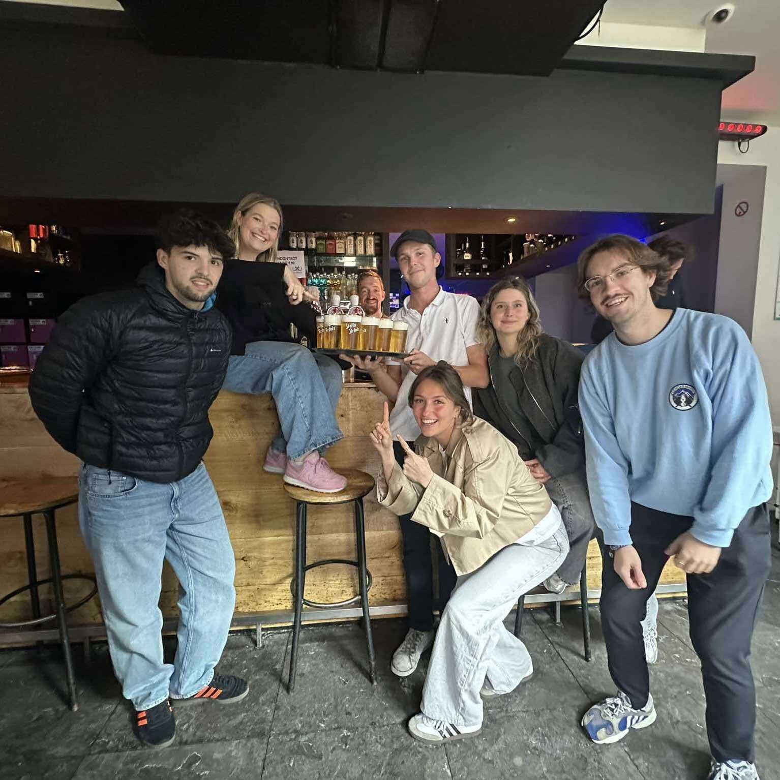 Six people standing in front of the bar, one holding a tray of beers, and one person behind the bar in Café De Vagant.