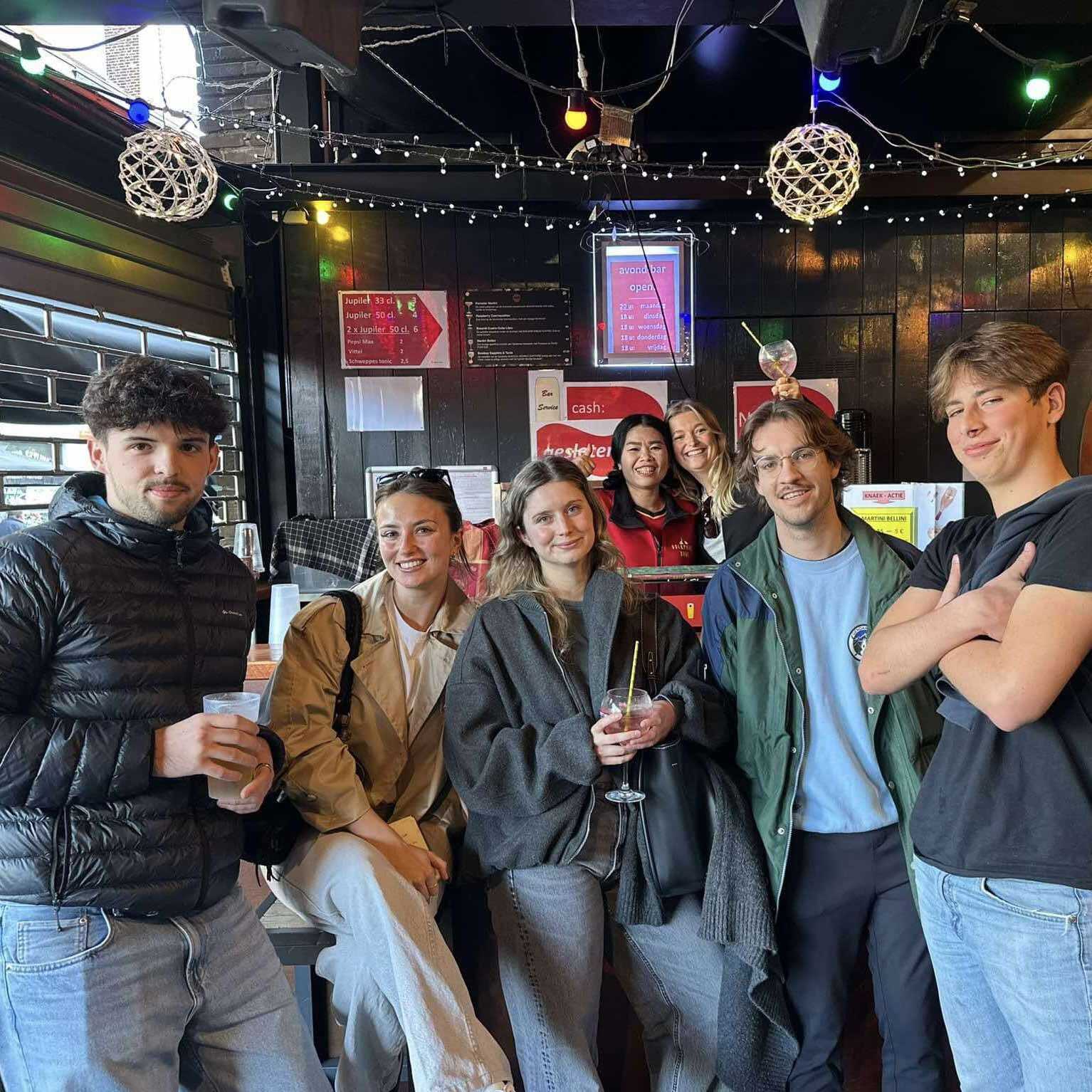 Group of friends posing and smiling together in a cozy, warmly lit bar with colorful lights and ceiling decorations, some holding drinks.