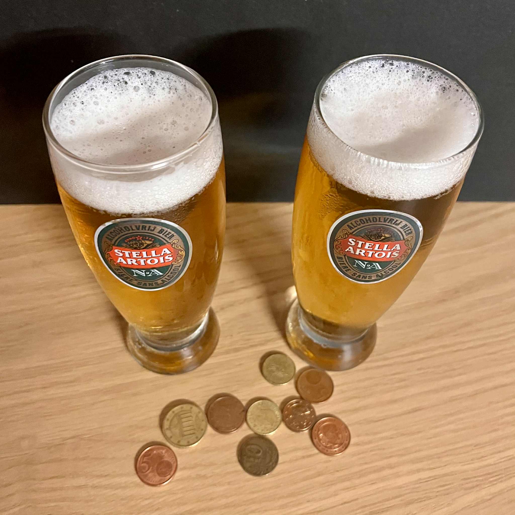 Two glasses of blond beer on a wooden table with coins in front of them.