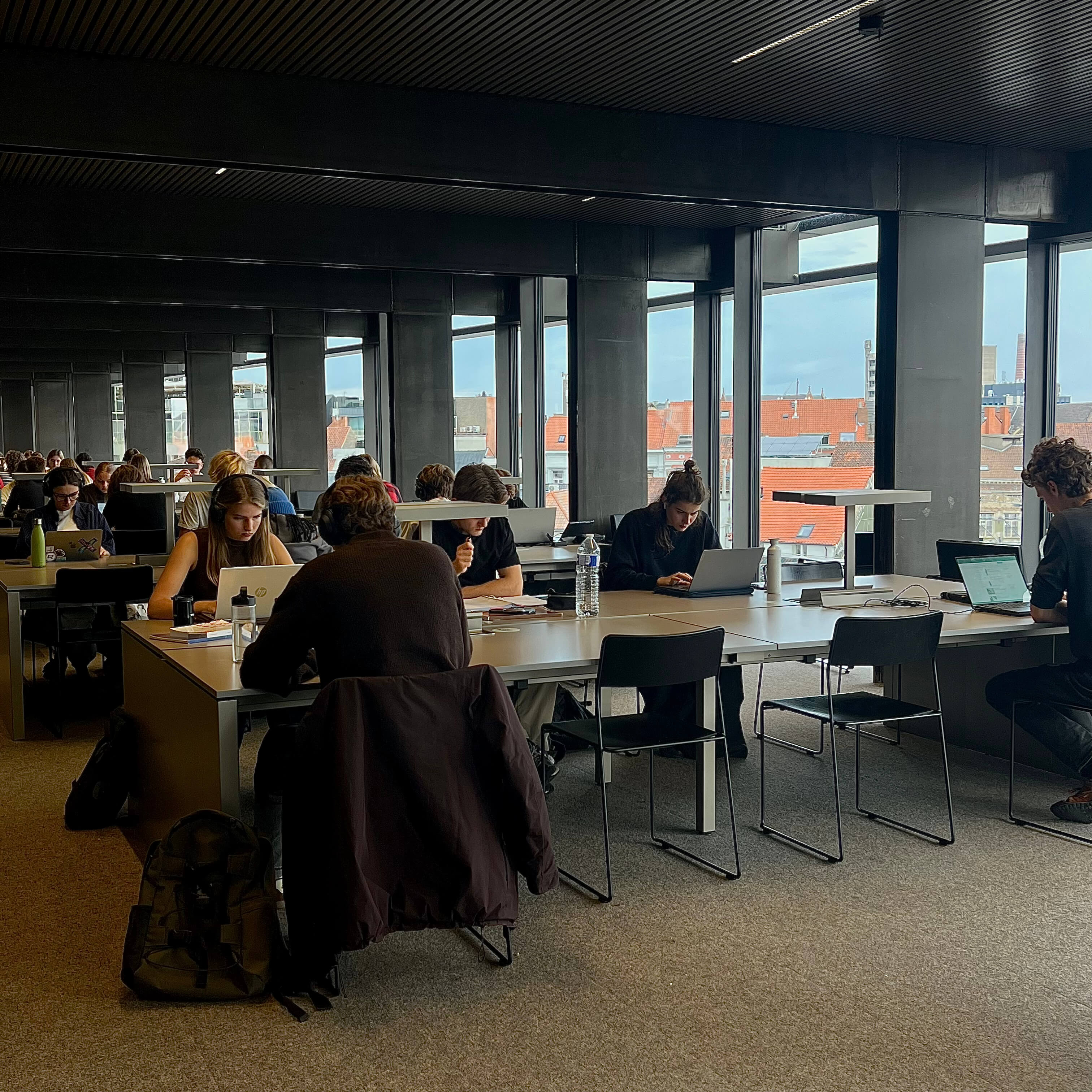 A busy, modern  study hall in Ghent's De Krook Library filled with students working on laptops at long desks, featuring large windows with a view of the cityscape.
