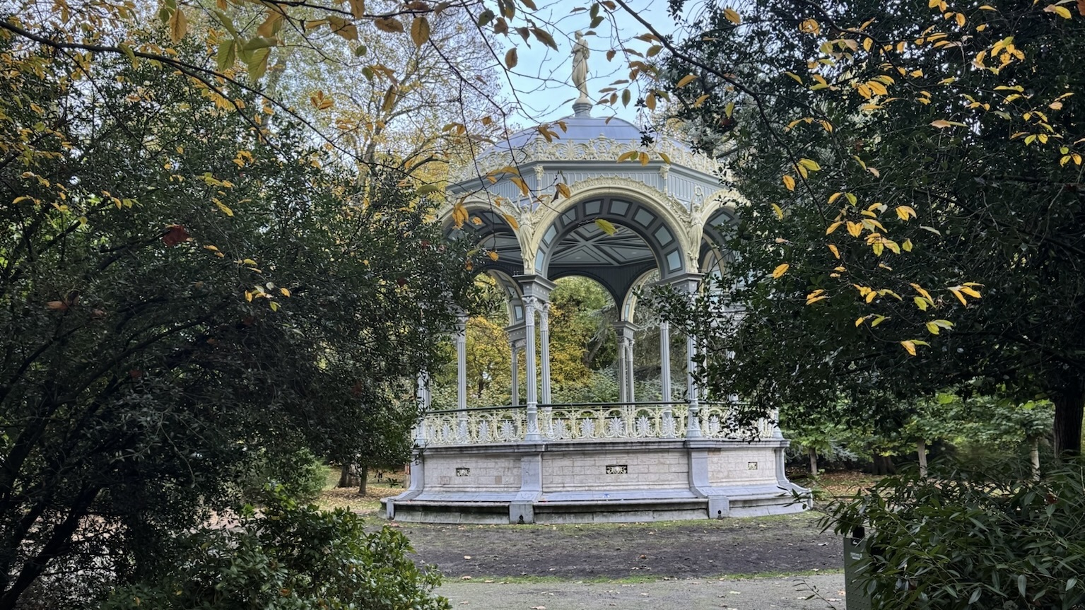 An elegant gazebo with ornate arches and a statue on top, surrounded by dense greenery and trees with autumn leaves.
