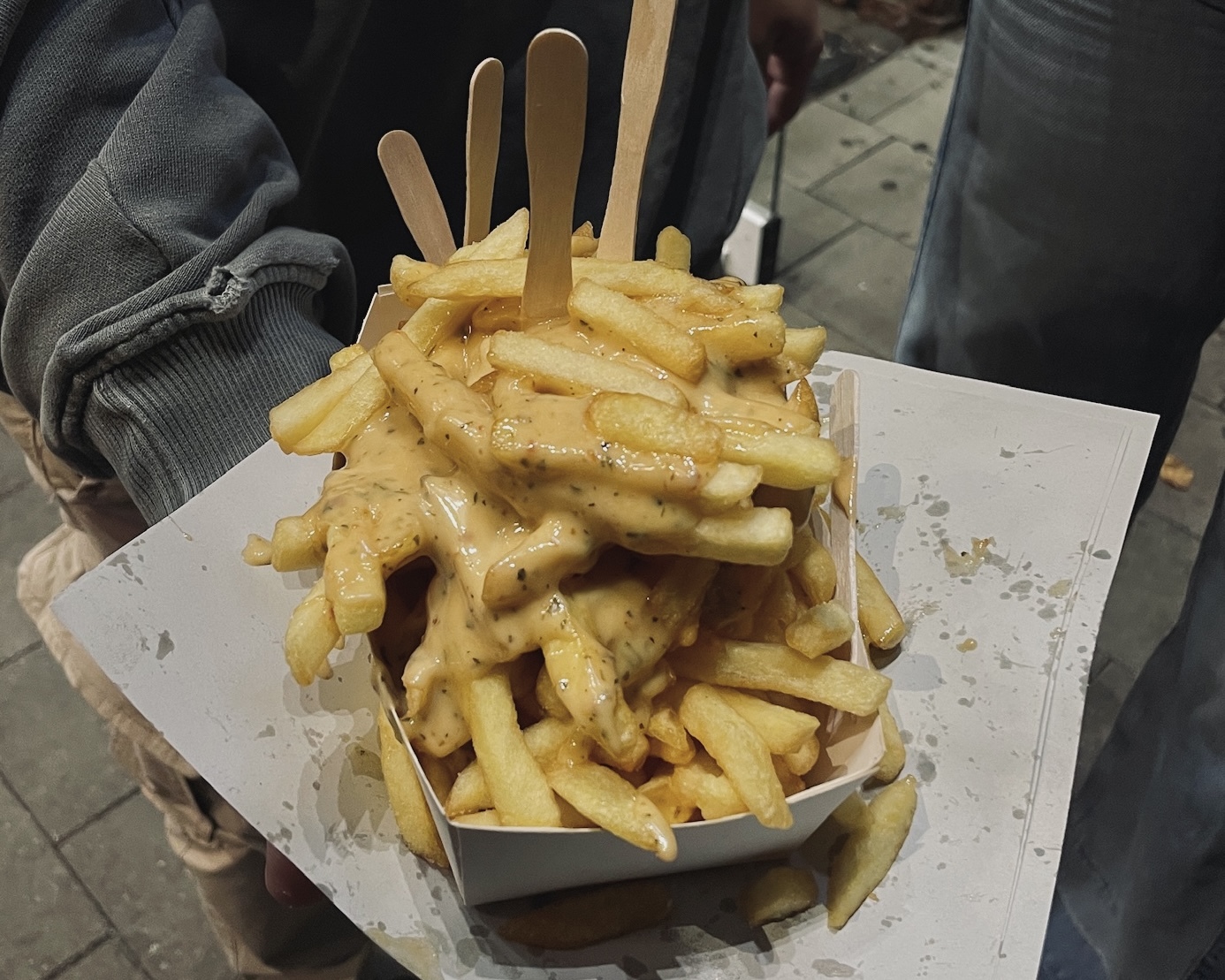 A close-up shot of a paper tray overflowing with golden French fries heavily topped with a creamy, light brown sauce, possibly a seasoned cheese sauce or an elaborate mayonnaise. Several small wooden forks are sticking out of the fries.