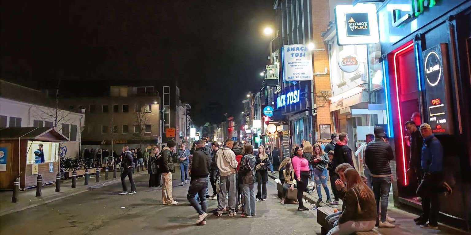 Night scene in Overpoortstraat, Ghent, with groups of people socializing outside bars and snack shops. The street is lively with young adults, mostly students, enjoying the nightlife atmosphere under bright neon signs and streetlights.